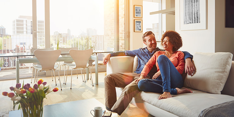 A mixed-race couple relaxing on the couch in an apartment with bright natural lighting.