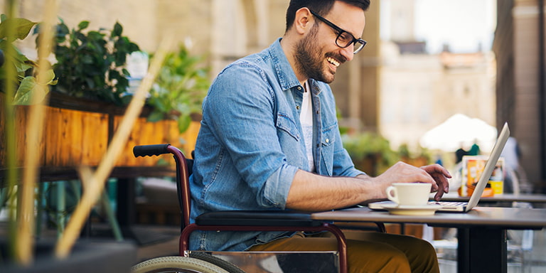 A man in a wheelchair sitting outside at a restaurant and having a coffee whilst working on his laptop.