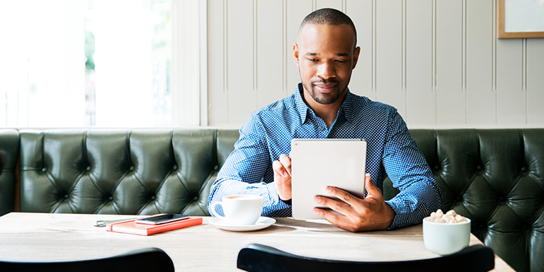 A man sitting at a cafe and having a cup of coffee whilst busy on his tablet.