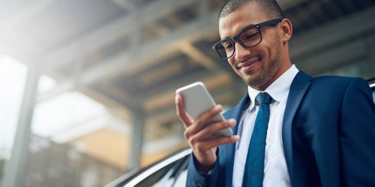 A man in a blue suit, white shirt and blue tie standing next to his car looking at his phone while smiling as his personalised car and home insurance quote was just sent through.