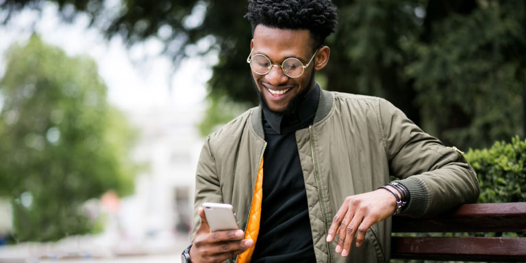Young man sitting on a bench outdoors, smiling while looking at his cell phone, reading up on Momentum’s Evolve medical aid option.