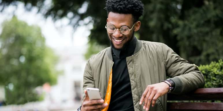 Young man sitting on a bench outdoors, smiling while looking at his cell phone, reading up on Momentum’s Evolve medical aid option.