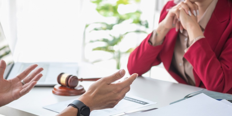 An experienced executor, sitting at her office desk, next to a gavel, with legal documentation, discussing a deceased’s Will with a beneficiary.