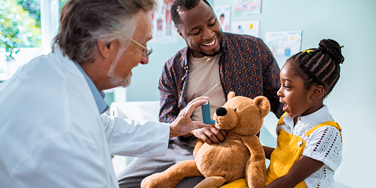 A father and daughter at a doctor's consultation, sitting on the medical bed, while the doctor demonstrates how to administer an asthma pump on a teddy bear.