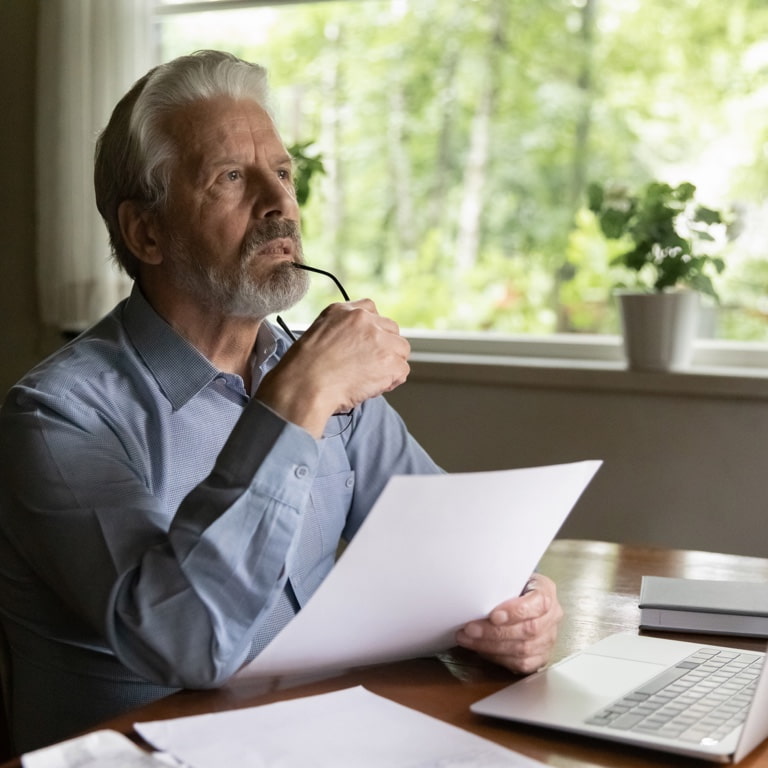 An older man with greying hair, holding a paper in front of his laptop deep in thought. He is holding his glasses in his right hand while resting them on his chin.