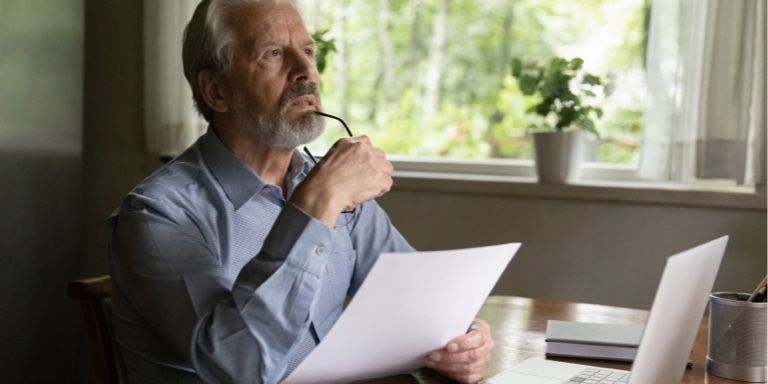 An older man with greying hair, holding a paper in front of his laptop deep in thought. He is holding his glasses in his right hand while resting them on his chin.