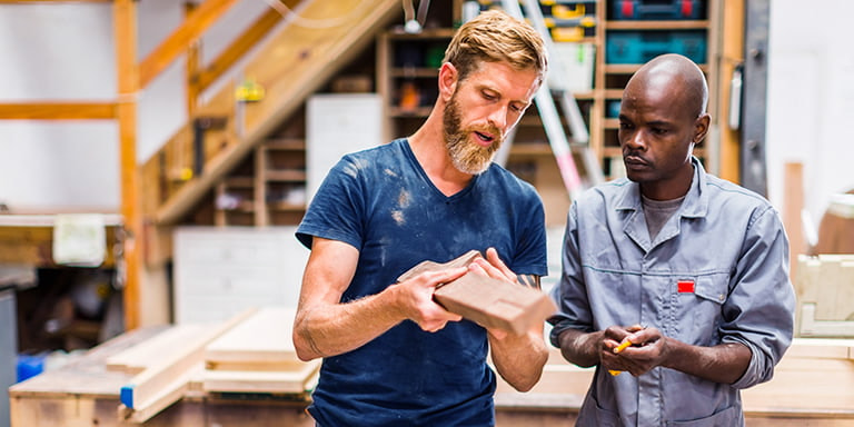 An employer holding a piece of wood in a carpentry workshop and explaining to one of his employee what needs to be done. 