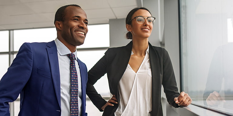 Work colleagues, a man in a navy suit and a woman in a black blazer and white blouse, smile and look out a window.