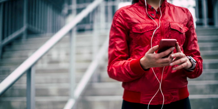 Leading a healthy lifestyle, an athletic lady in a red sporting gear, holding her cell phone with earphones in her ears with stairs in the background.