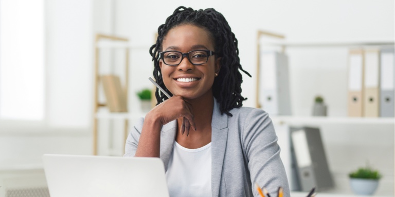 An African woman in front of her laptop at the office, confidently smiling ahead after updating beneficiaries on her retirement fund.