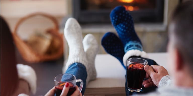 A couple sit in front of a fire with their sock-covered feet on the coffee table, drinking a warm drink.