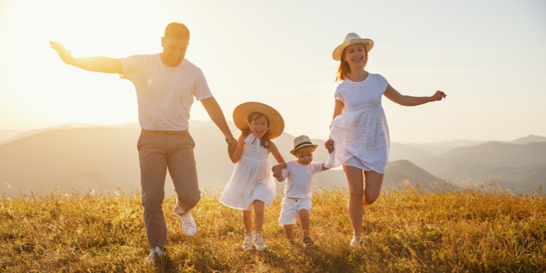 A family of four, mother and father holding their two children’s hands as they run in an open field as the sun sets.