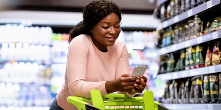 Women in the shopping isle of a grocery store, leaning on her trolly while looking at her cell phone.