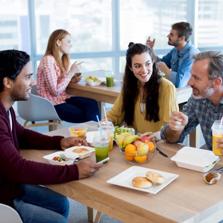 A young male and female sitting in a restaurant eating a healthy breakfast of fruit, smoothies, and bagel while conversing with one another.