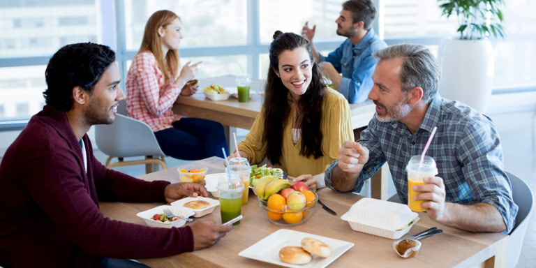 Three colleagues, two men and a woman, smiling and talking while they sit in the canteen and enjoy their lunch together.