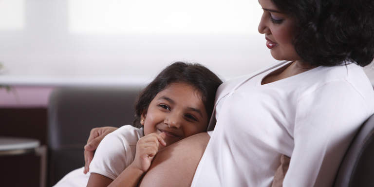 Young pregnant mom sits with her little daughter who is leaning on her tummy in the doctor's waiting room.