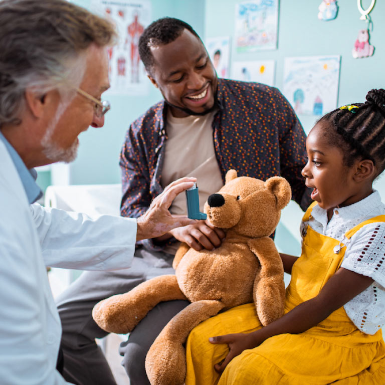 A father and daughter at a doctor's consultation, sitting on the medical bed, while the doctor demonstrates how to administer an asthma pump on a teddy bear.