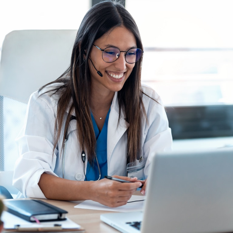 Young women's specialist sitting in an office working on her laptop, smiling while consulting with a patient online through Momentum’s Hello Doctor.
