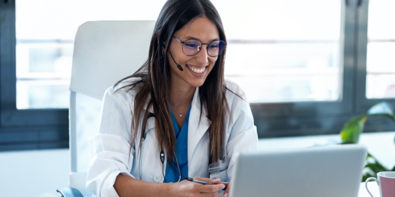 Young women's specialist sitting in an office working on her laptop, smiling while consulting with a patient online through Momentum’s Hello Doctor.