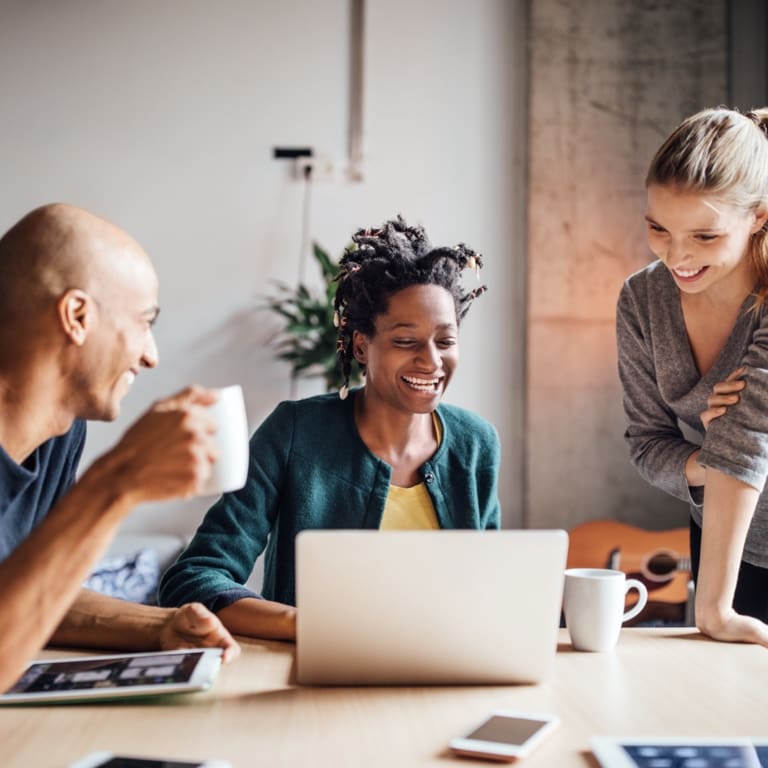Two women an a male colleague, with a coffee mug in his hand, having a meeting around a table, looking at the laptop screen of the woman in the middle.