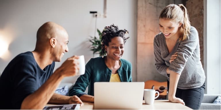 Two women and a male colleague, with a coffee mug in his hand, looking at the laptop screen of the woman in the middle.