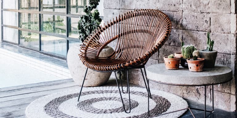 Cane chair on a grey and white coloured round carpet, next to a concrete table with metal legs that has a range of cacti plants displayed on it.