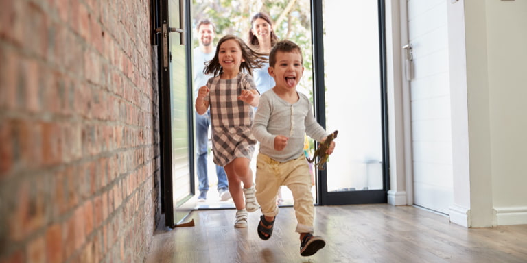 A couple standing outside of the door of their new home, watching their two toddlers running inside. 