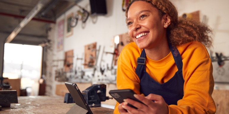 Woman smiling while in front of workshop counter with a cell phone in her hand.