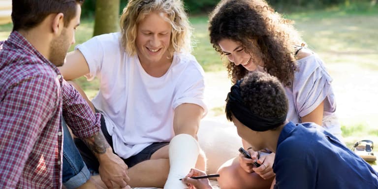 A family sit under a big tree in the park while the youngest male draws on one of the male's cast on his arm.