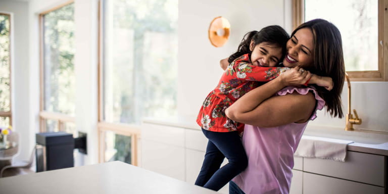 A young mum hugs her daughter excitedly in the kitchen of their home.