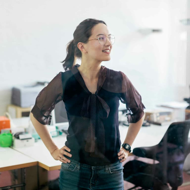 A young lady with a big smile on her face is wearing round spectacles and a black blouse with sheer sleeves standing in an office with her hands on her hips.