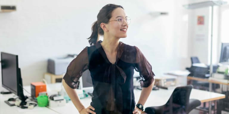 A young lady with a big smile on her face is wearing round spectacles and a black blouse with sheer sleeves standing in an office with her hands on her hips.