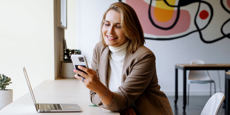 Young working professional woman seated at her desk in front of her laptop looking at her cell phone.