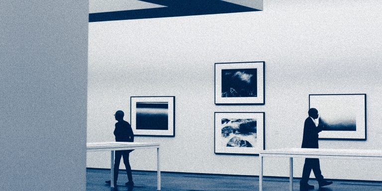 Two people walking in a modern art gallery with framed abstract photographs on the wall and white display tables in the foreground.