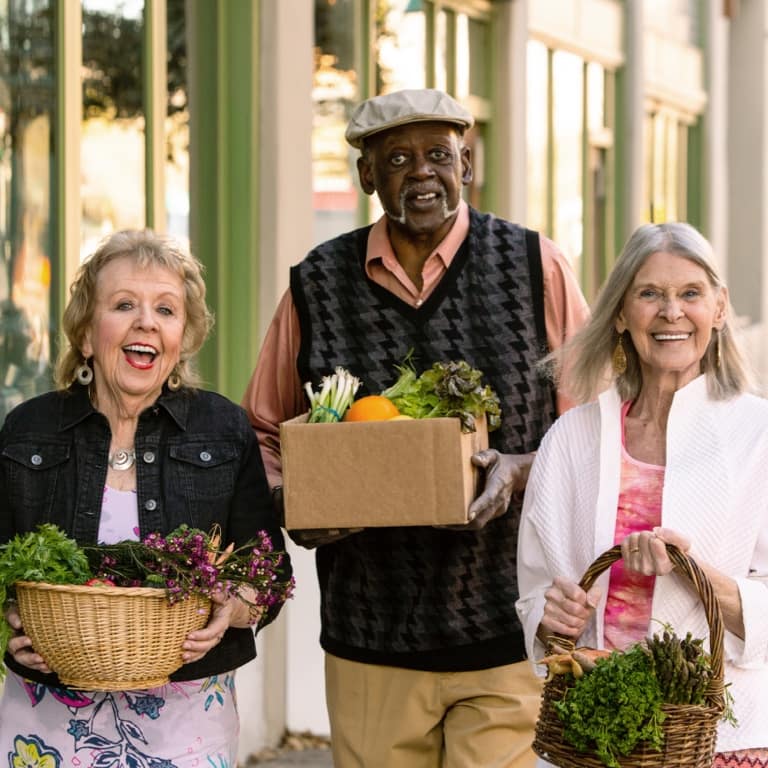 A retired, elderly African man walking alongside two white retired women, all carrying vegetable-filled baskets.