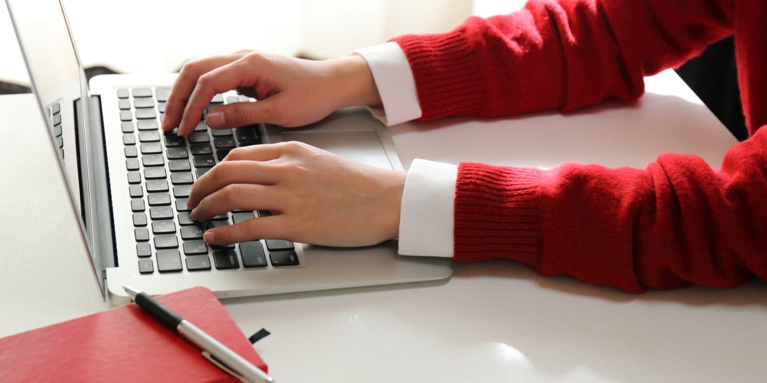A close-up of two hands typing on a laptop with a red notebook next to the laptop.