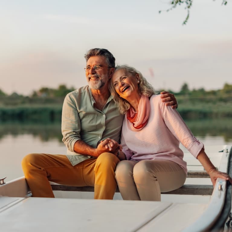 An older couple sits on a bench by a lake, holding hands and smiling. Trees and calm water create a peaceful, natural environment.