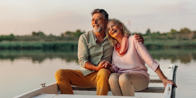 An older couple sits on a bench by a lake, holding hands and smiling. Trees and calm water create a peaceful, natural environment.