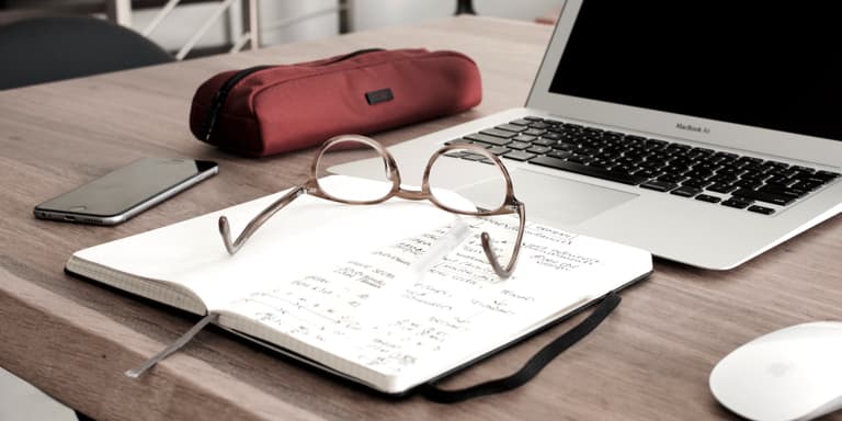 An open diary with the owner's glasses on top of it, on a table, by a laptop, cell phone and pencil case.