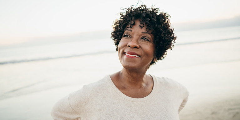 A woman with short, black, curly hair standing by the seashore with her hands on her hips, smiling.