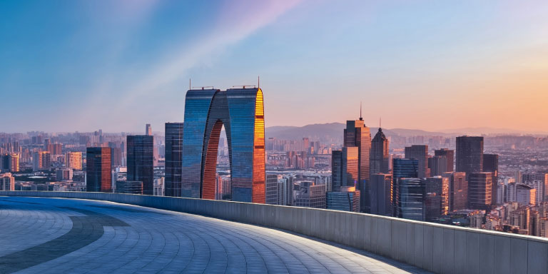 Cityscape view at sunset featuring modern skyscrapers, including a distinctive arch-shaped building, seen from a curved rooftop or terrace with a clear sky in the background.