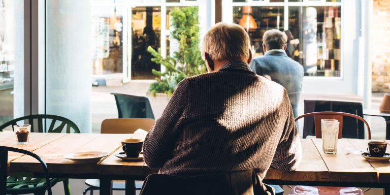Elderly man sitting at a table in café drinking a cup of coffee and looking out the window with a view of the shops in front of him.