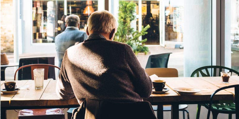 Elderly man sitting at a table in café drinking a cup of coffee and looking out the window with a view of the shops in front of him.