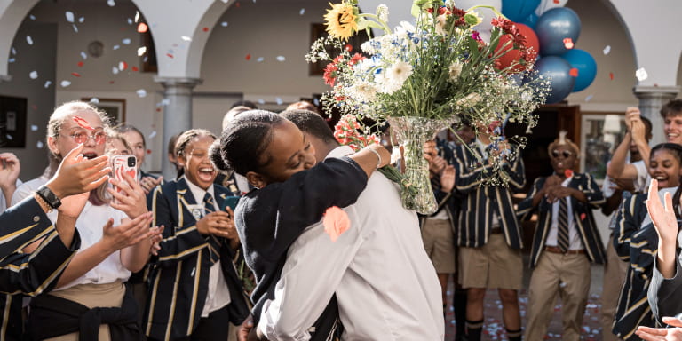 A scene from Momentum’s latest brand TV commercial featuring a heartfelt matric dance proposal. The girl, holding a vase of flowers, shares a warm hug with her date while a group of excited students in the background capture the moment on their cell phones
