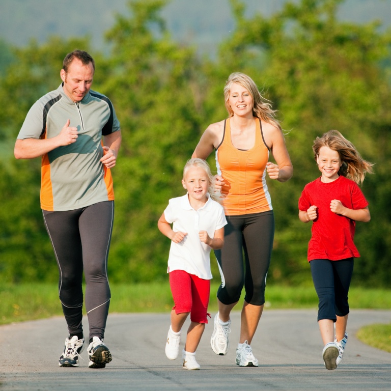 An energetic family of 4 - a mother, father, and 2 young children jogging on a country path.