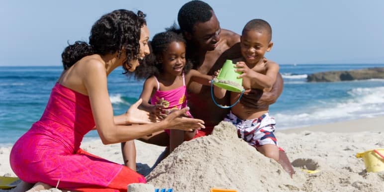 Mother and father happily playing on the beach with their two children in the sand building a sandcastle, near the ocean.
