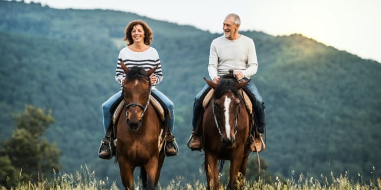 An elderly woman and man, happily riding horses outdoors with mountains in the background.