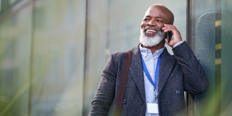A middle aged African male professional speaking on his mobile phone, standing outside of an office building wearing a coat.