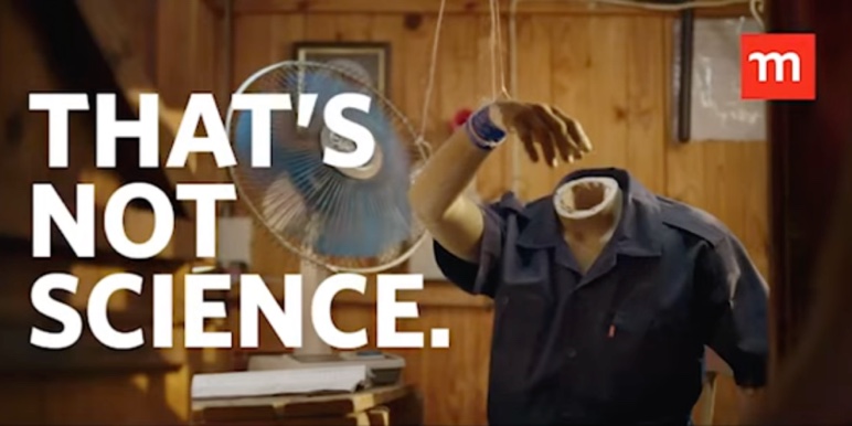 A mannequin dressed in a blue overall seated in an outdoor building. A man stands next to the mannequin holding the mannequin’s head.