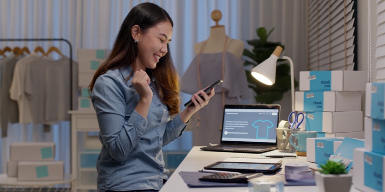 Fashion designer sitting at her work desk looking excitedly at her phone.